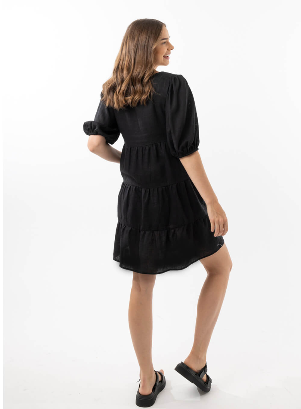 The Eleanor mini dress in black features a flowing layered a line shape with draping linen/viscose fabric, puff 3/4 sleeves with elastic cuff which can be pushed up to the elbow, a dropped v shape neckline with a square finish.